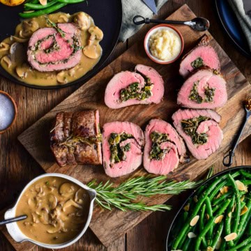 A sliced stuffed roast beef surrounded with a bowl of gravy, a dish of green beans, potatoes, horseradish, salt, napkins and cutlery.