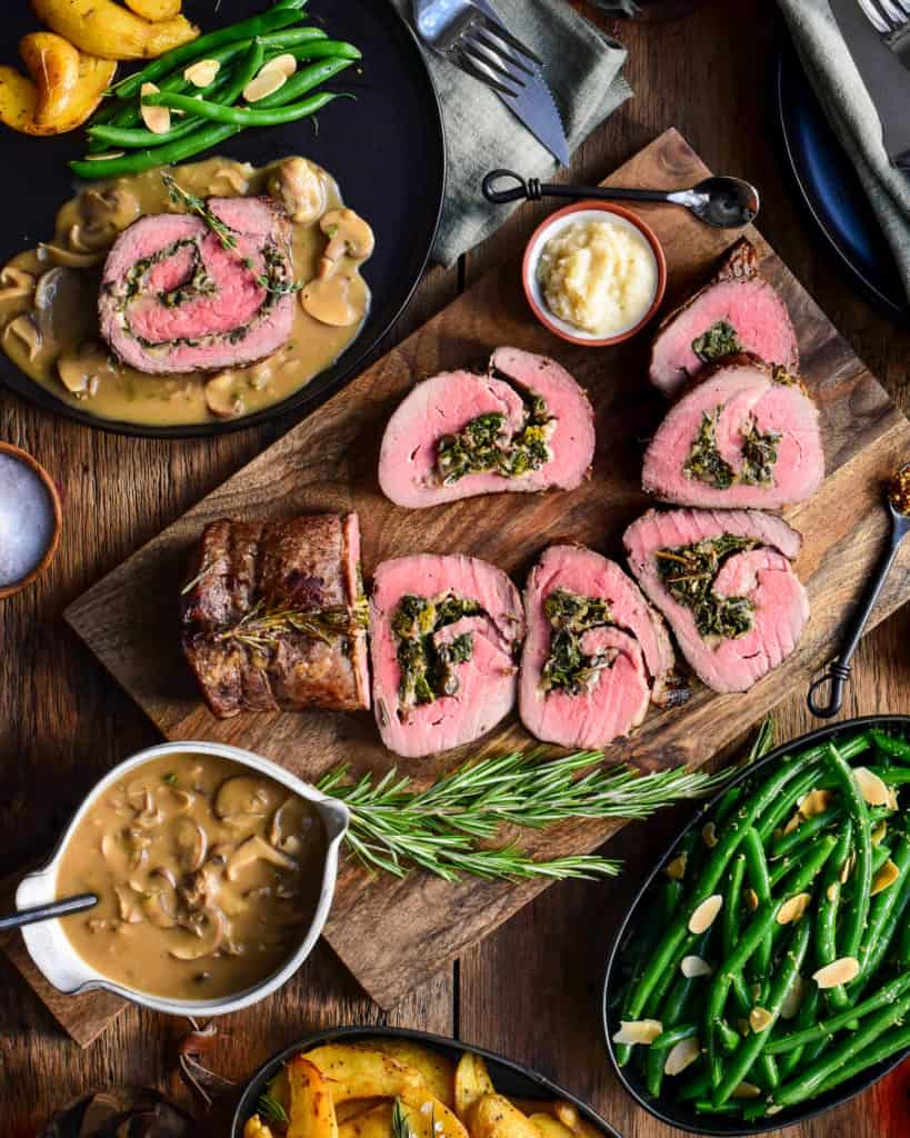 A sliced stuffed roast beef surrounded with a bowl of gravy, a dish of green beans, potatoes, horseradish, salt, napkins and cutlery.