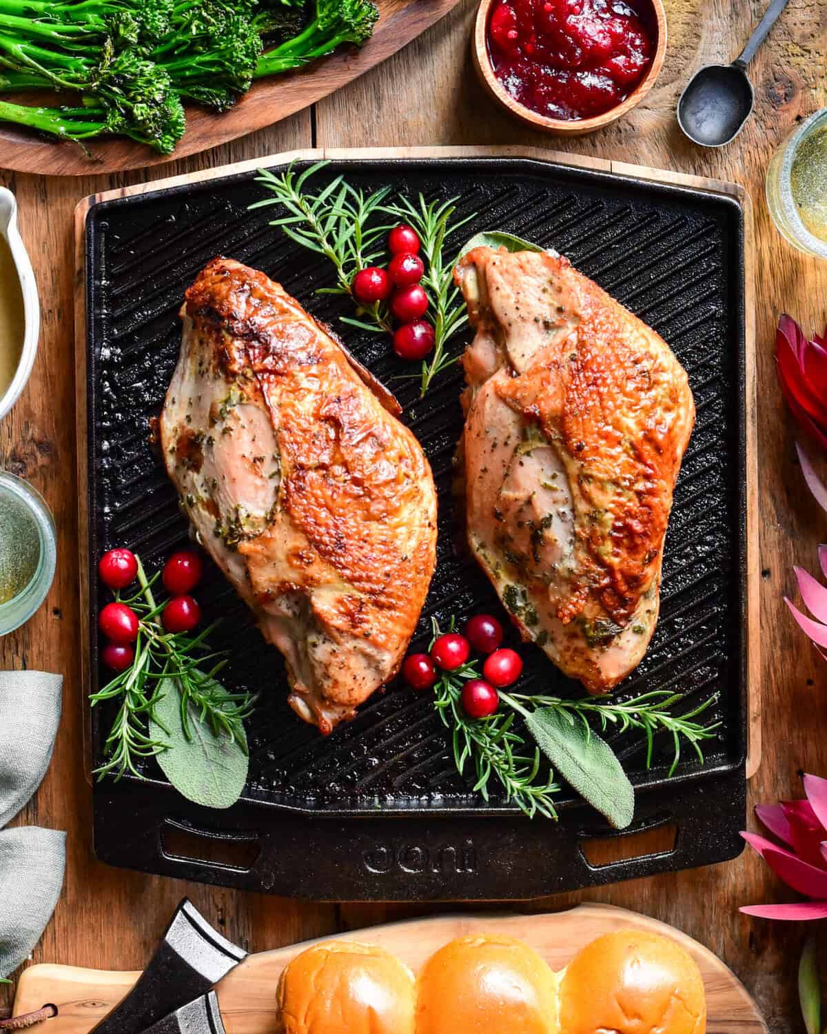 A top down image of two cooked turkey breasts on an Ooni grizzler skillet with herbs and cranberries. Broccolini, carrots, gravy, buns, cranberry sauce, wine glasses and florals surround this skillet.
