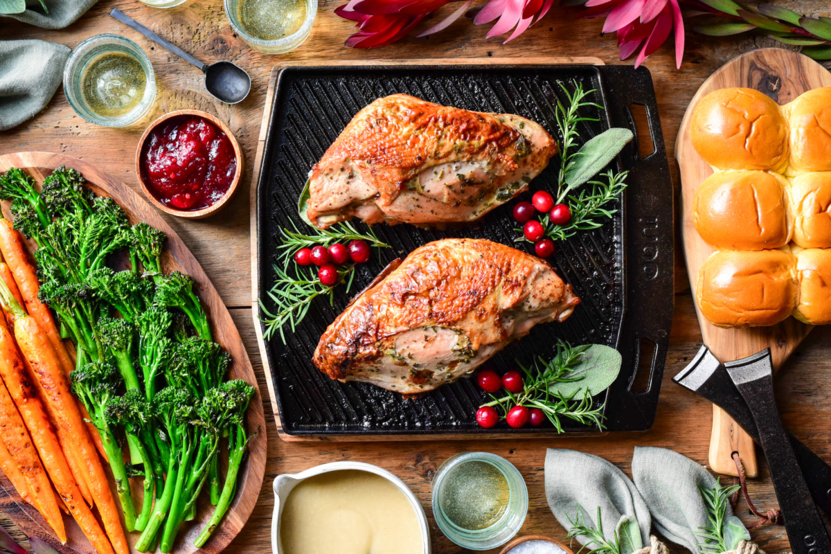 A top down image of two cooked turkey breasts on an Ooni grizzler skillet with herbs and cranberries. Broccolini, carrots, gravy, buns, cranberry sauce, wine glasses and florals surround this skillet.