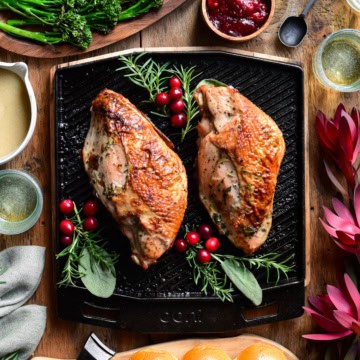 A top down image of two cooked turkey breasts on a skillet with herbs and cranberries. Broccolini, carrots, gravy, buns, cranberry sauce, wine glasses and florals surround this skillet.