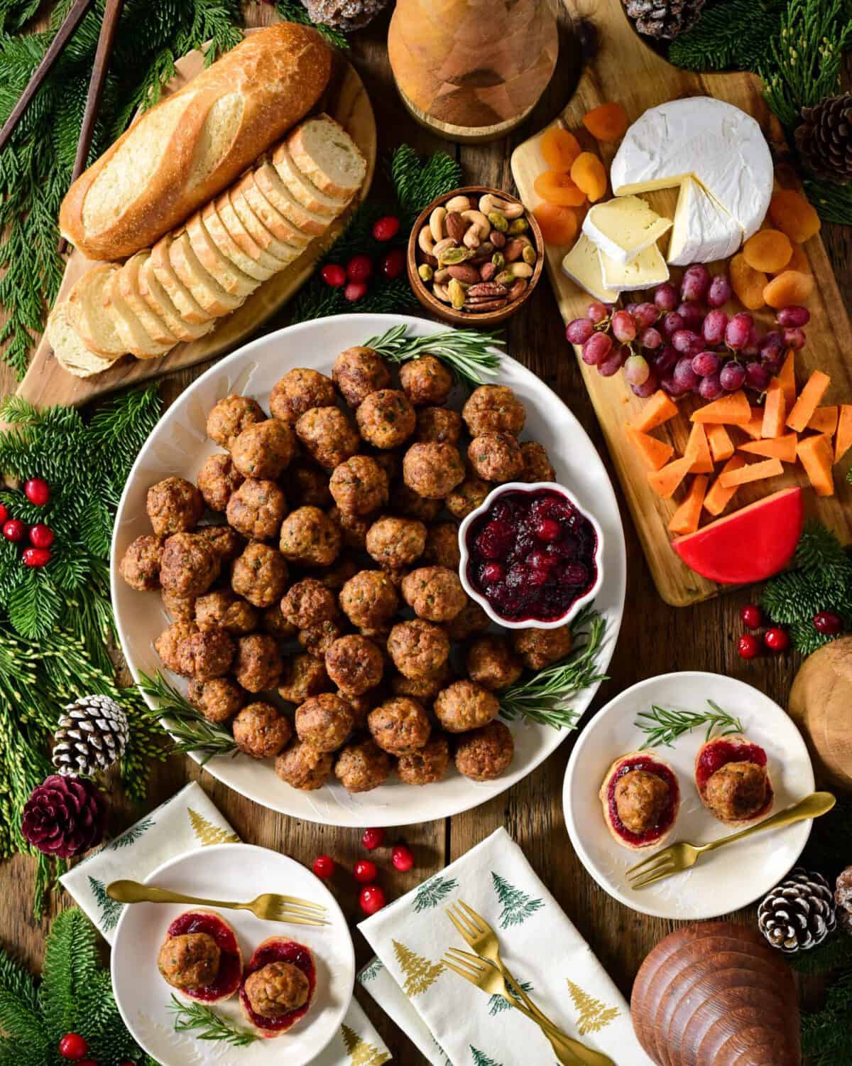 A top down image of a round platter filled with meatballs and bowl of cranberry sauce. A sliced baguette, a cheese board, plates and festive decoration surround this platter.