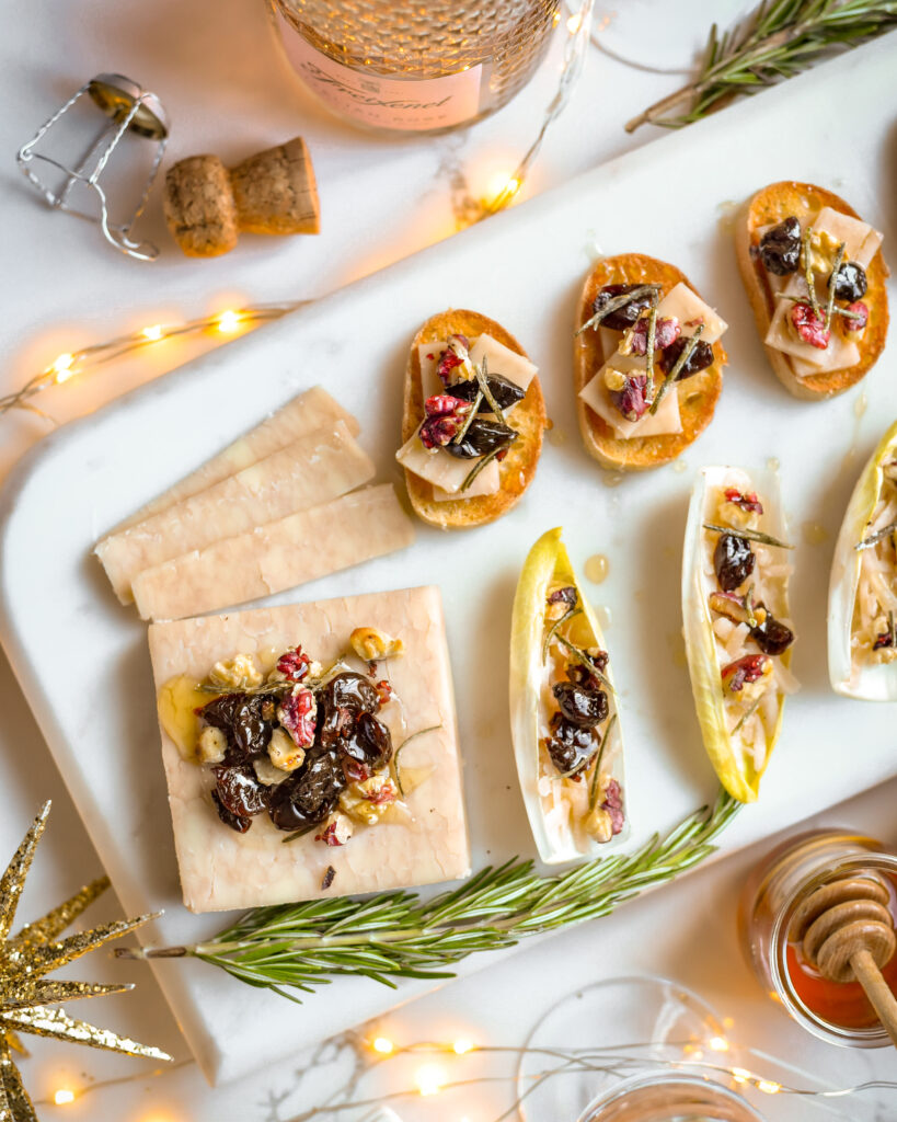 A marble tray of cheese crostinis and endive boats with a block of cheese all topped with walnuts, honey, dried cherries and rosemary.