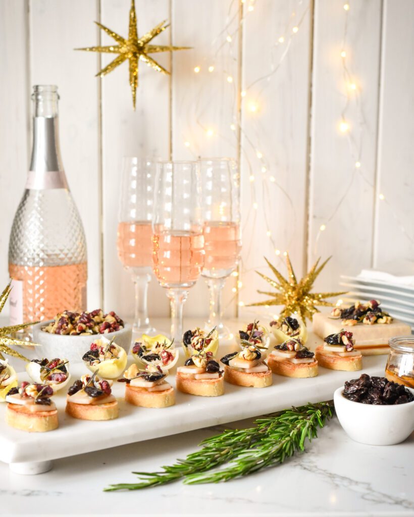 An image of Red-Wine Cheddar Crostini & Endive Boats with a bottle of pink champagne, glasses, a bowl of dried cherries and walnuts.