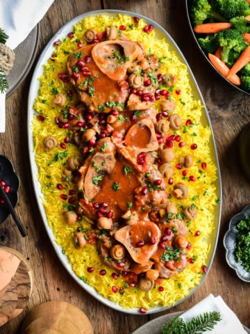 An overhead image of an oval platter with veal shanks and gravy over rice. Toppings and vegetables surround this platter.