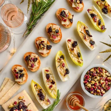A marble tray of seven cheese crostinis, and endive boats with a block of cheese all topped with walnuts, honey, dried cherries and rosemary. Surrounded by glasses of pink Champagne, rosemary, honey and twinkle lights.