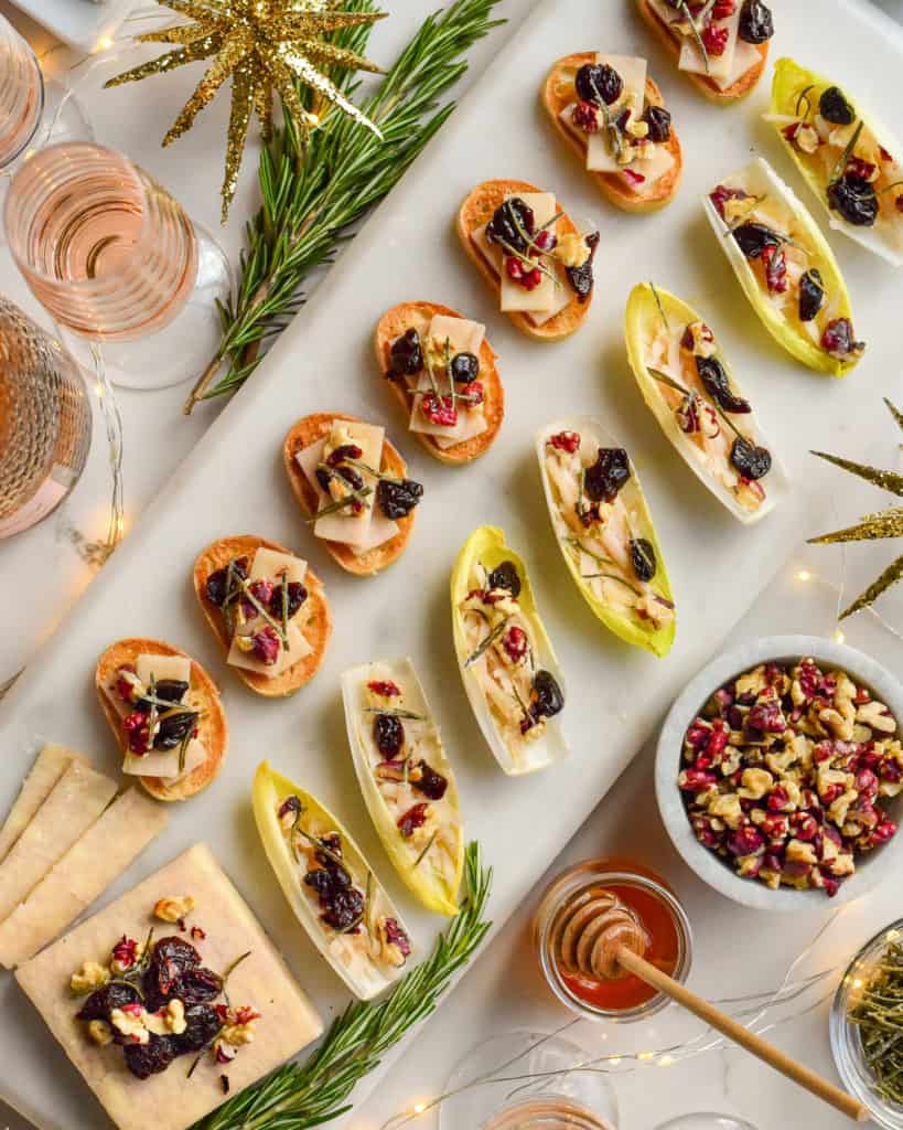 A marble tray of seven cheese crostinis, and endive boats with a block of cheese all topped with walnuts, honey, dried cherries and rosemary. Surrounded by glasses of pink Champagne, rosemary, honey and twinkle lights.