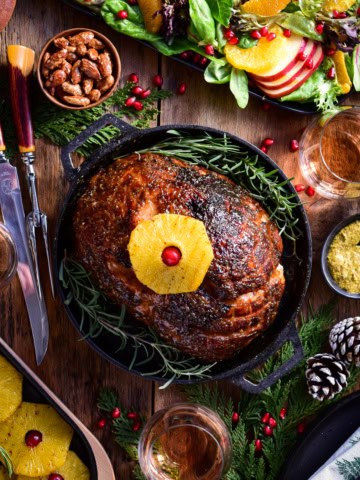 An image of a baked ham in a cast iron skillet with a pineapple on top. There is also winter salad, more baked pineapples, a carving knife set and holiday sprigs on the table.