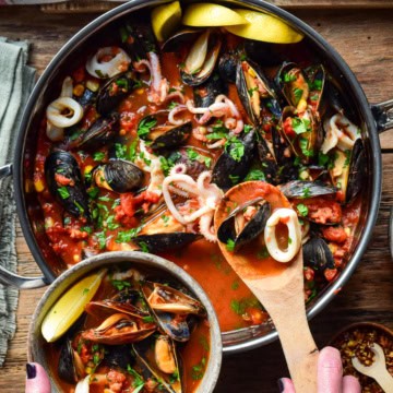 A top down image of large pot of mussels and squid in a tomato broth. A portion is being spooned out into a bowl.
