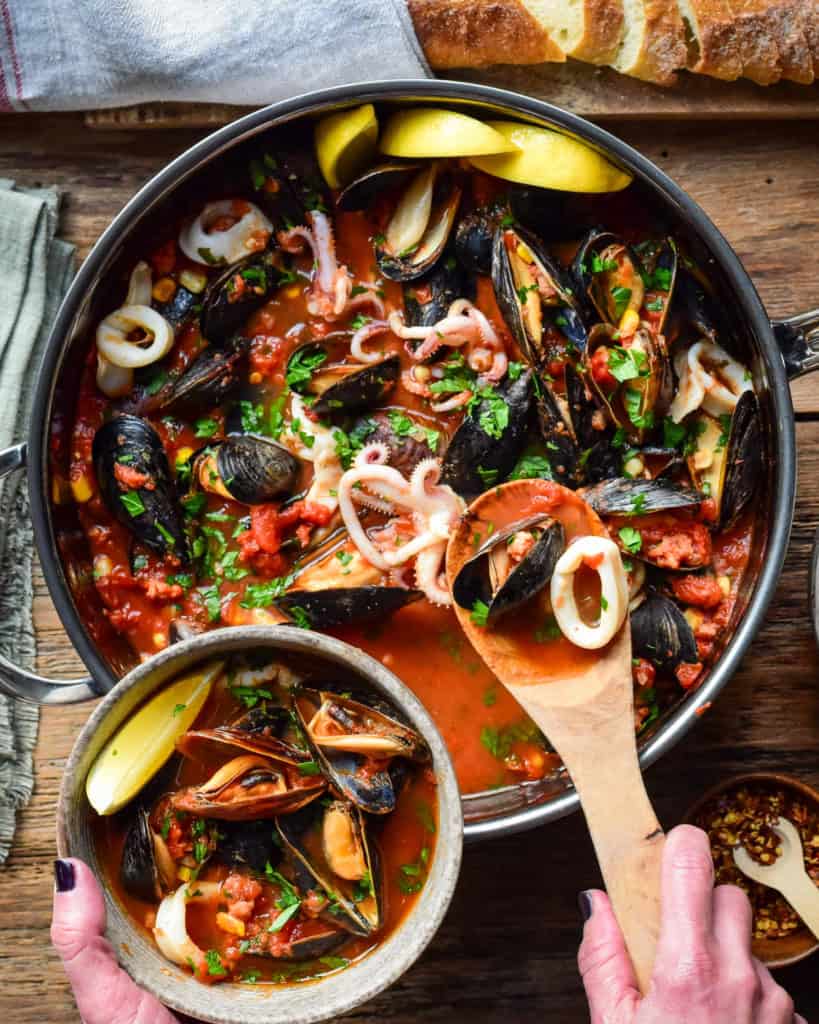 A top down image of large pot of mussels and squid in a tomato broth. A portion is being spooned out into a bowl.