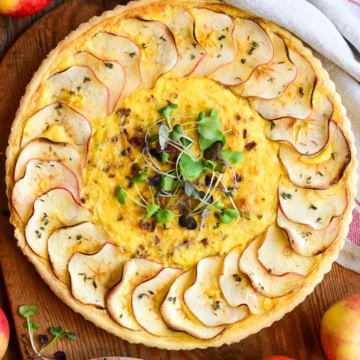 An apple, leek, bacon quiche with apple slices around the perimeter and garnished with mircogreens.