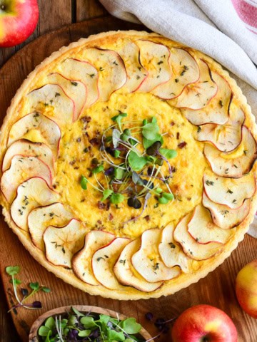 An apple, leek, bacon quiche with apple slices around the perimeter and garnished with mircogreens.