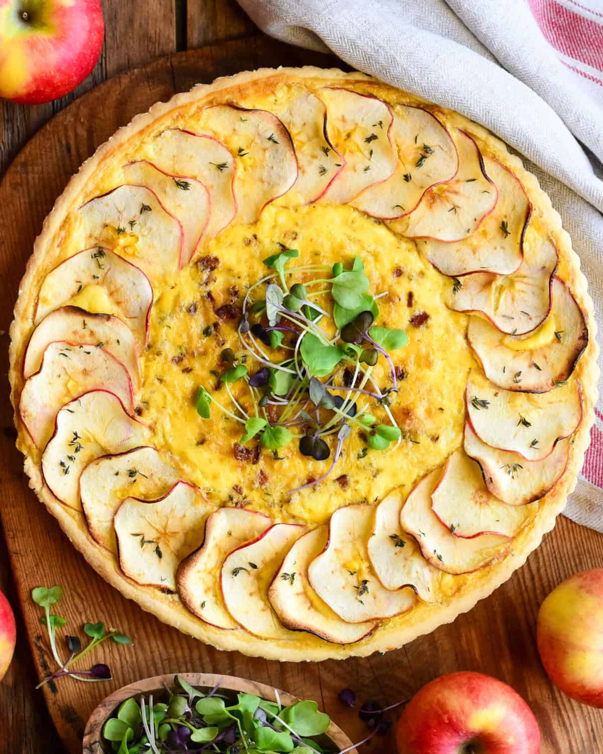 An apple, leek, bacon quiche with apple slices around the perimeter and garnished with mircogreens.