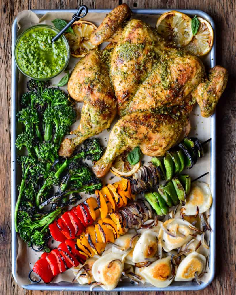 A sheet pan with a grilled pesto spatchcocked chicken, charred broccolini, grilled peppers & onions and fried pierogi with onions.