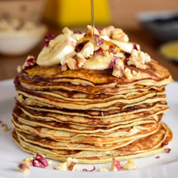 A large stack of two ingredient banana pancakes with bananas and nuts on the top with syrup.