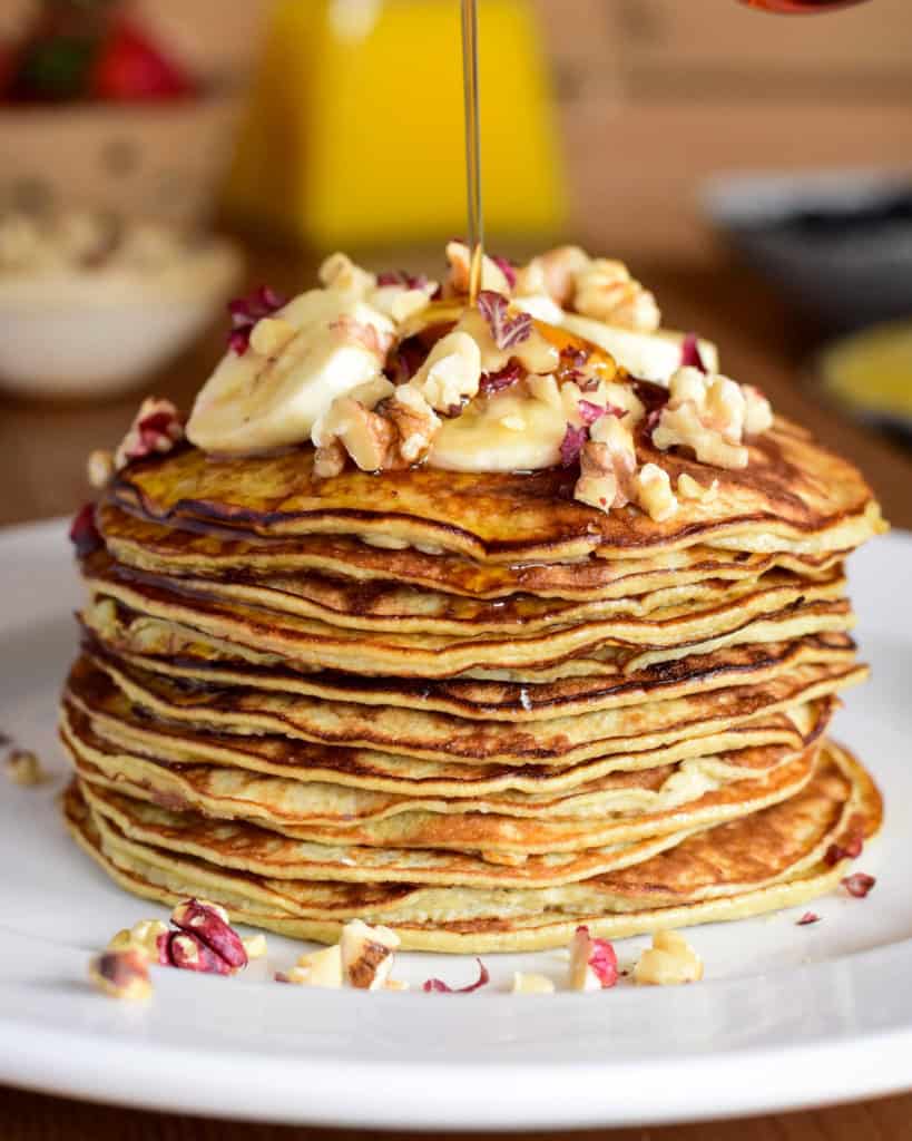 A large stack of two ingredient banana pancakes with bananas and nuts on the top with syrup.