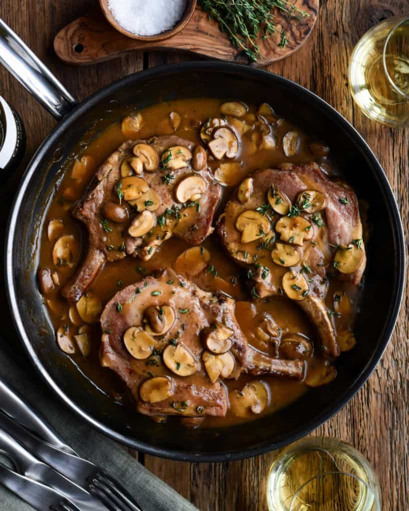 A skillet with three bone-in pork chops nestled in a luscious mushroom sauce.