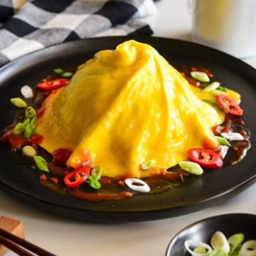 A domed and swirled tornado omelette (Omurice) over a pile of rice (not shown) with sauce and toppings on the base.