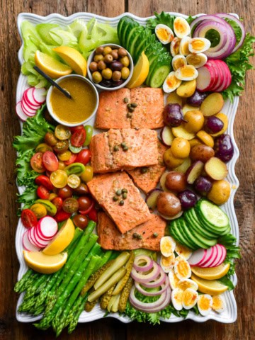 A large platter of a Niçoise Salad with Salmon. Five pieces of salmon are centered in the platter and are surrounded by various vegetables, eggs and pickles.