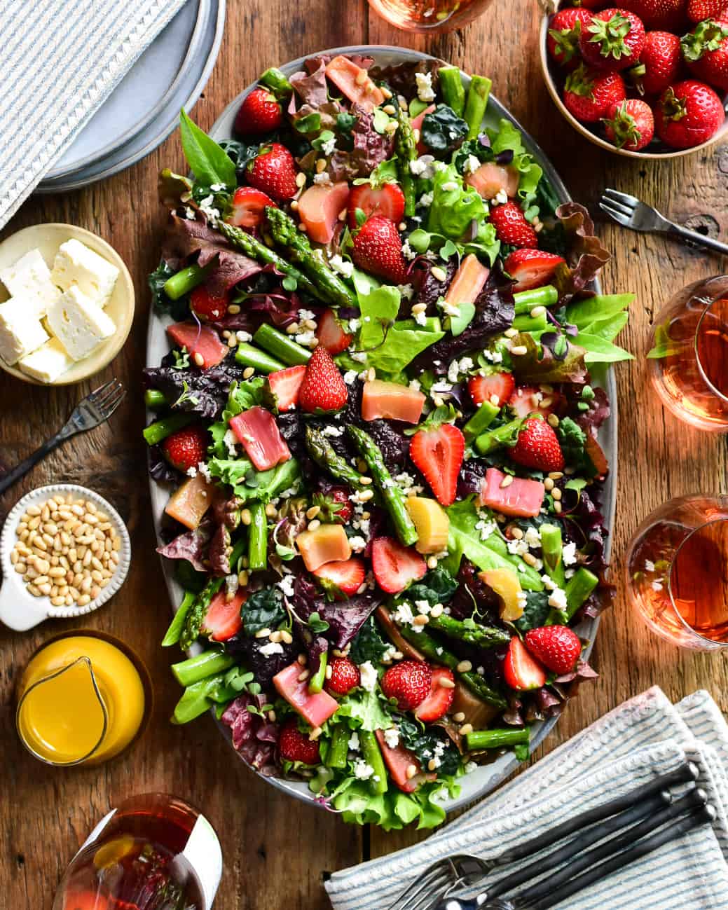 A top down image of a large Strawberry, Asparagus and Rhubarb Salad with dressing, feta, pine nuts and rose glasses surrounding it.