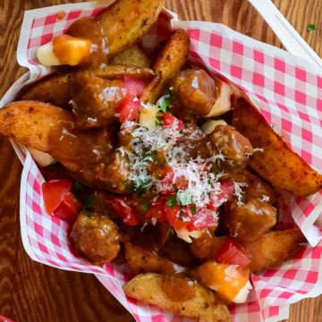 A top down view of poutine with meatballs. A red and white check snack bowl if filled with potato wedges and cheese curds covered in gravy and meatballs.
