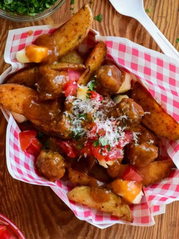 A top down view of poutine with meatballs. A red and white check snack bowl if filled with potato wedges and cheese curds covered in gravy and meatballs.