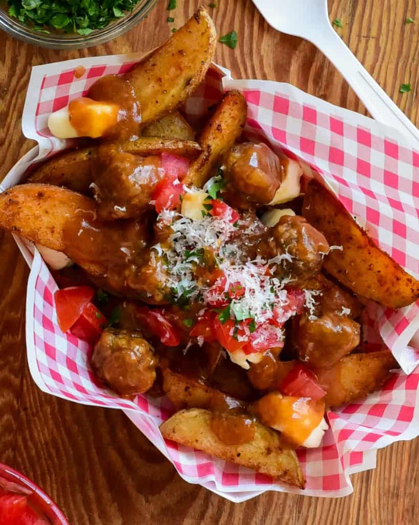 A top down view of poutine with meatballs. A red and white check snack bowl if filled with potato wedges and cheese curds covered in gravy and meatballs. 