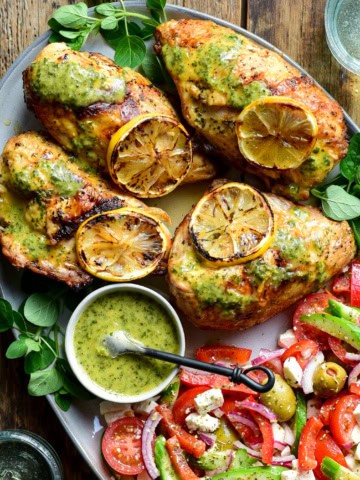 Four grilled and charred chicken breasts with charred lemon slices and served with a tomato salad.