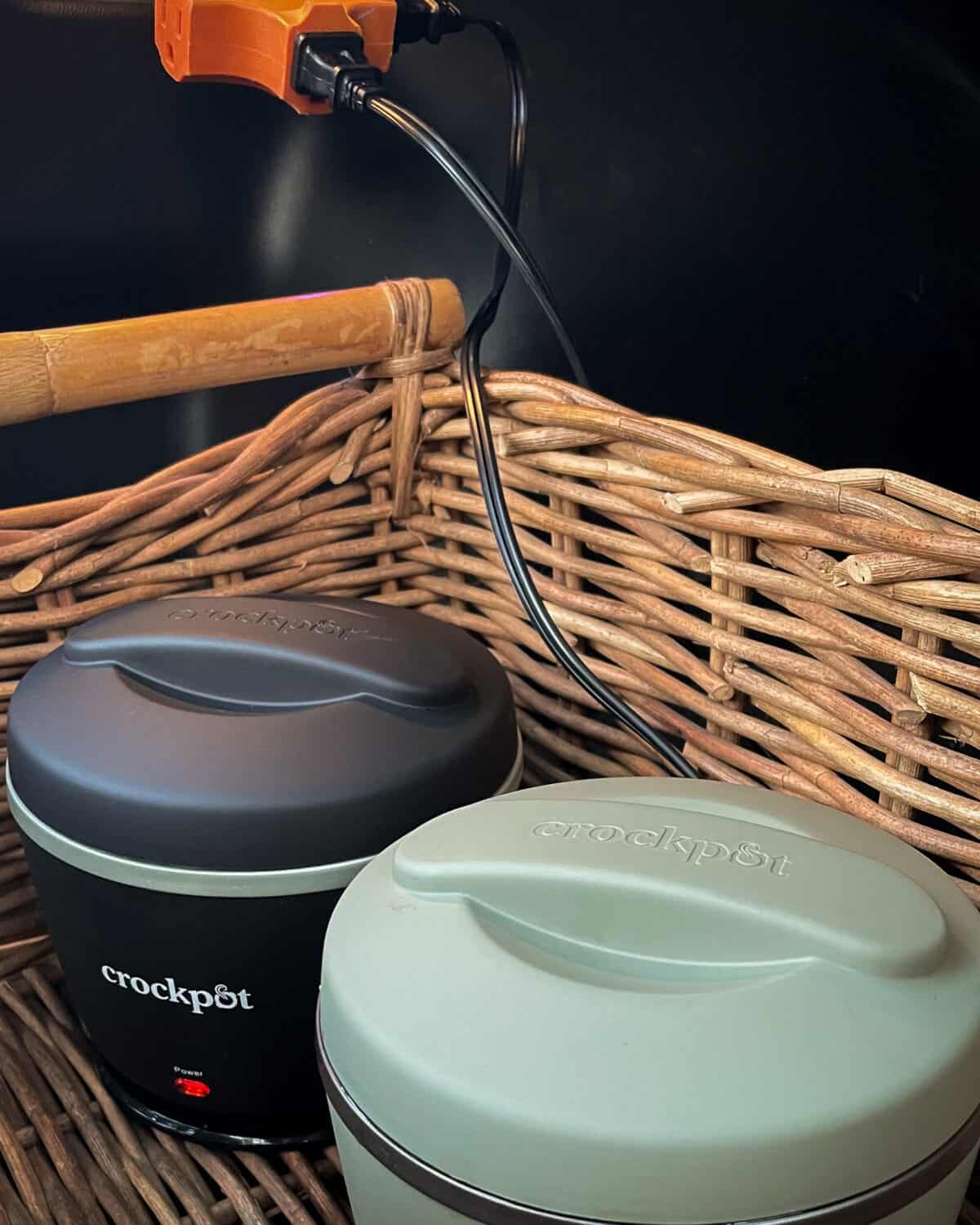 A black and a green mini lunch crock pot in a wooden basket getting charged in the drunk of a car.
