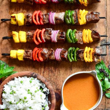 Four grilled beef skewers with alternating pieces of mango, peppers and onions on a board with a bowl of Thai curry sauce and a bowl of rice on a wooden board.