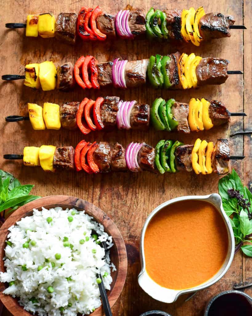 Four grilled beef skewers with alternating pieces of mango, peppers and onions on a board with a bowl of Thai curry sauce and a bowl of rice on a wooden board.