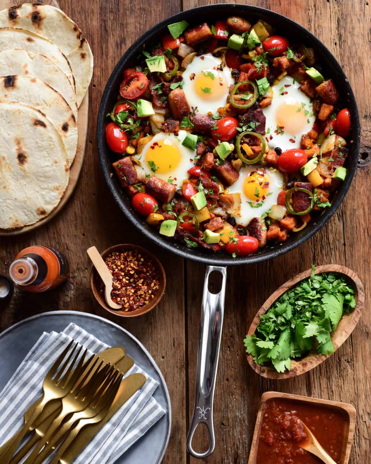 A one skillet hash with four eggs surrounded by plates, cutlery, hot sauce, cilantro and tortillas.