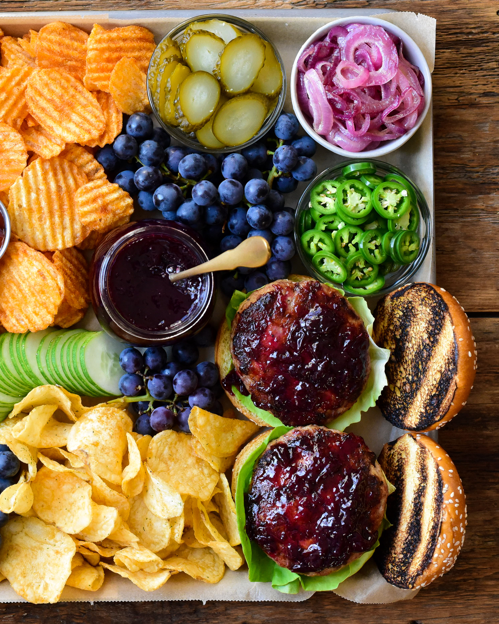 A close up of two turkey burgers with a grape jam in a tray with a jar of jam, chips, onions, jalapenos and grapes.