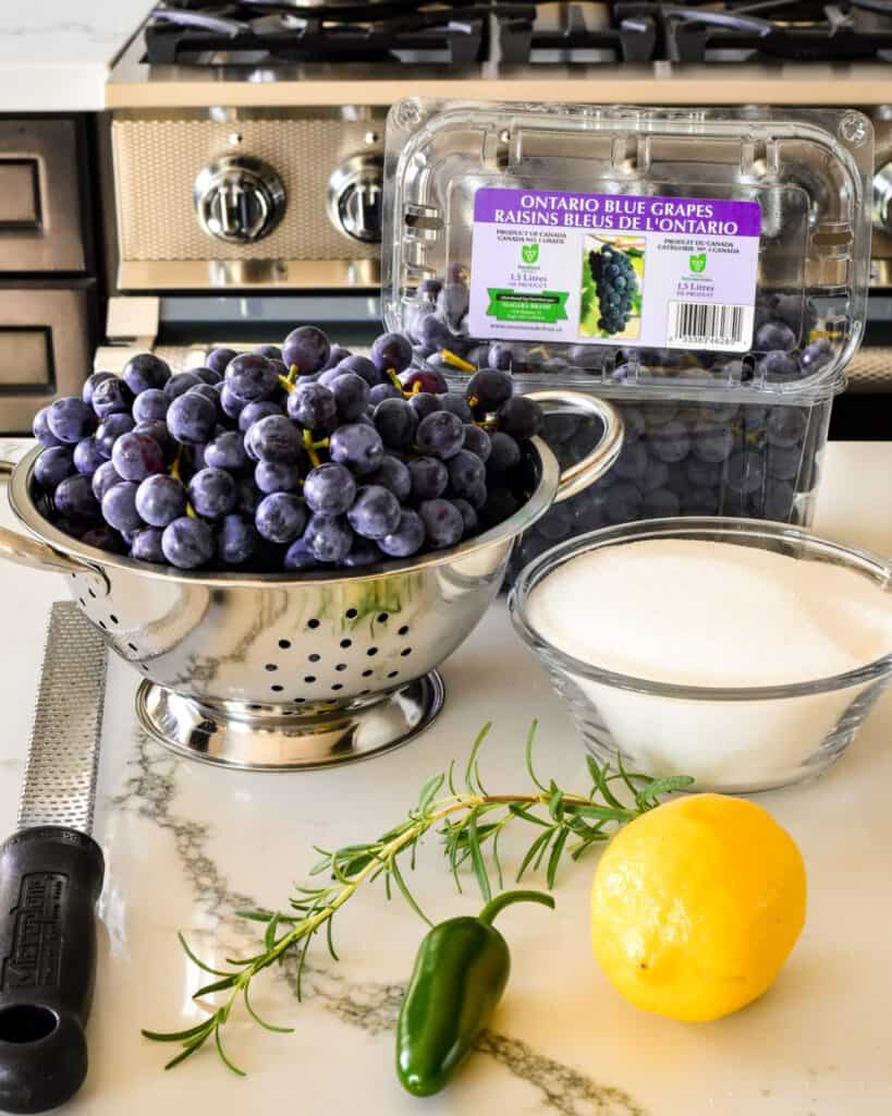 Ingredients for grape jelly without pectin are laid out: A bowl of blue grapes, sugar, rosemary, jalapeno and a lemon.