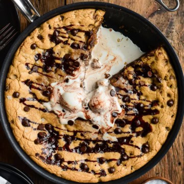 A top down image of a skillet chocolate chip cookie with a slice taken out and melted ice cream in the centre.