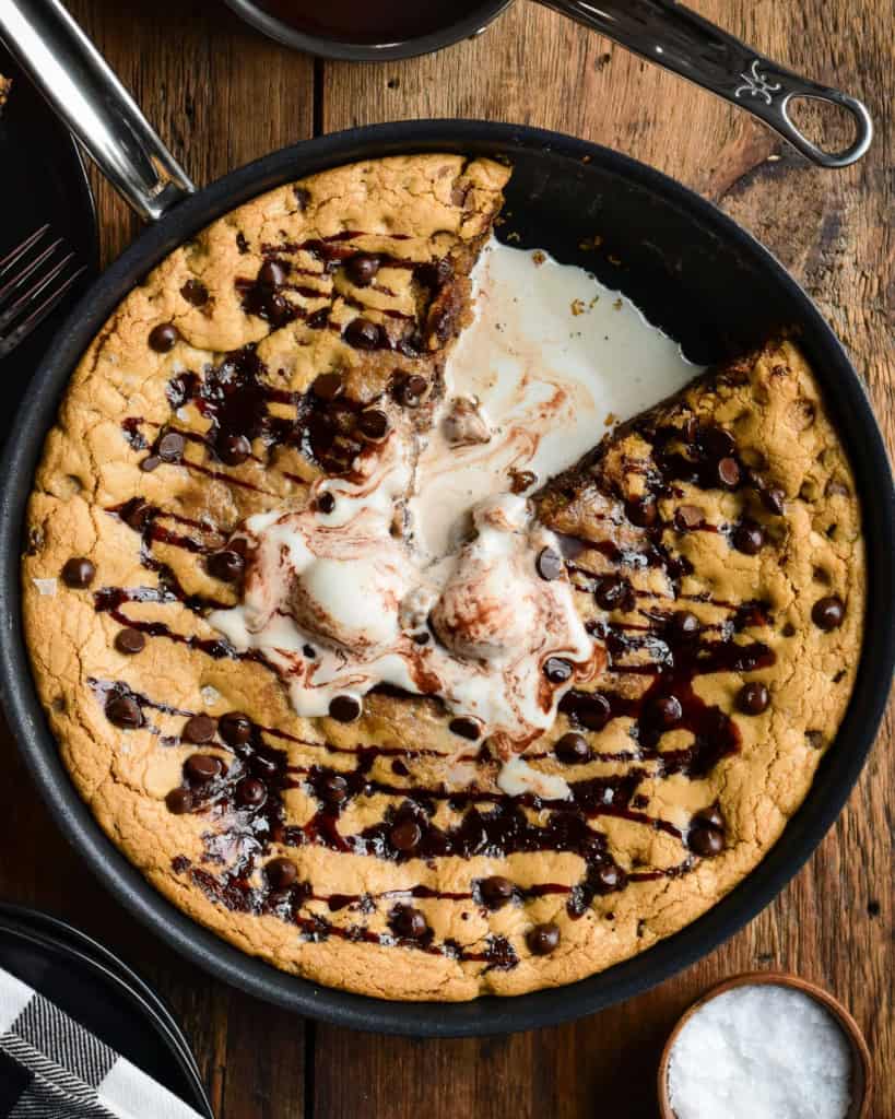 A top down image of a skillet chocolate chip cookie with a slice taken out and melted ice cream in the centre. 