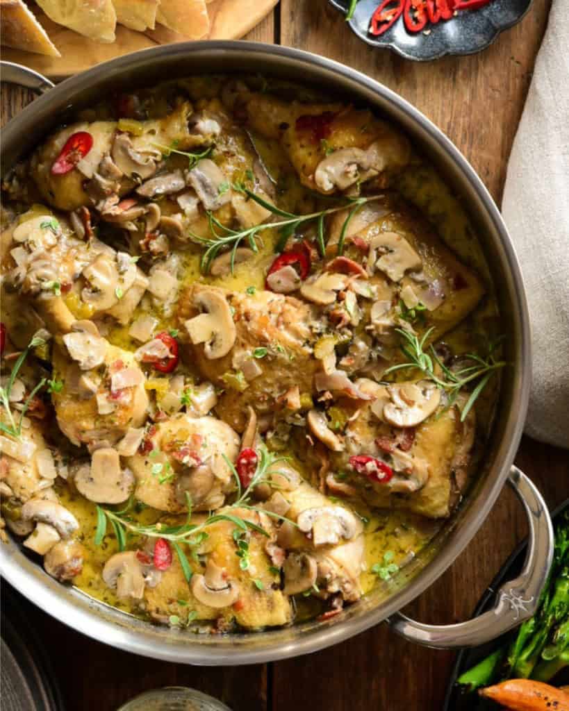 A close up of a finished chicken coq au vin blanc dish. The chicken is in a mushroom sauce with herbs and red peppers.