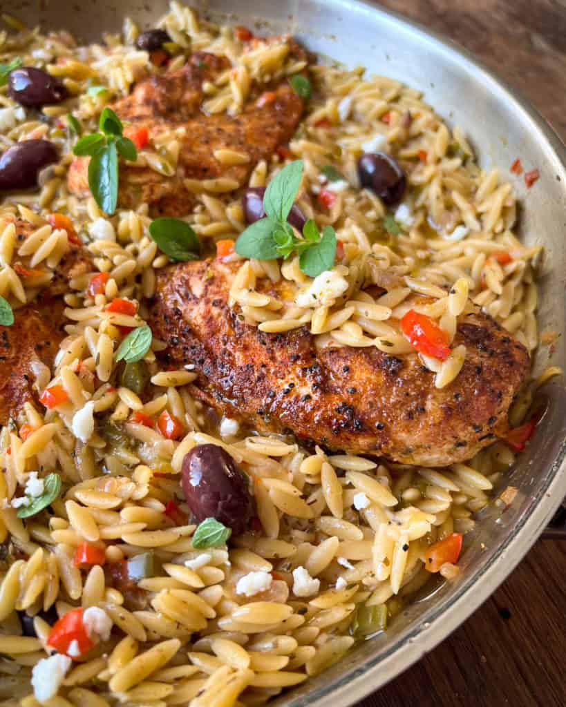 A skillet with chicken, orzo, olives and other ingredients.