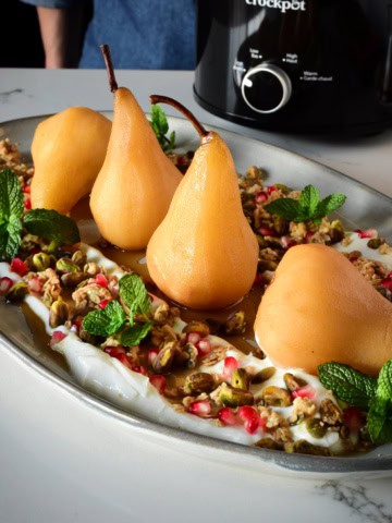 Four poached pears lined up on a platter surrounded with yogurt, nuts and granola with a black crockpot in the background.