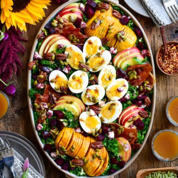 An oval platter with a Fall Harvest Salad with Apple Cider Vinaigrette consisting of boiled eggs, hasselback squash, apple slices and kale. Surrounded by sunflowers, dishes, glasses with apple cider and other salad toppings.