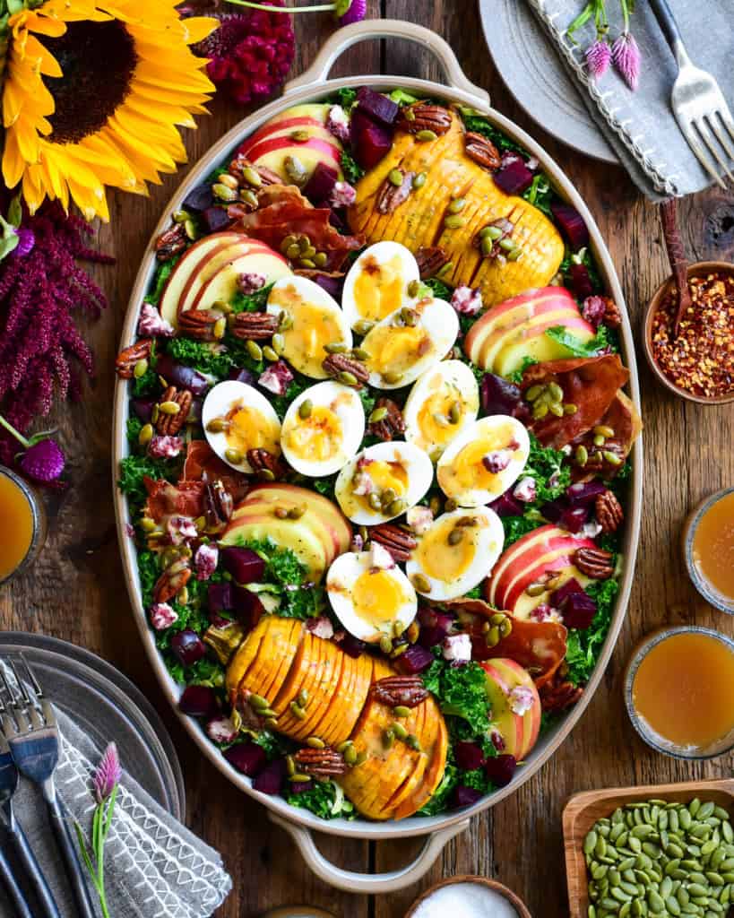 An oval platter with a Fall Harvest Salad with Apple Cider Vinaigrette consisting of boiled eggs, hasselback squash, apple slices and kale. Surrounded by sunflowers, dishes, glasses with apple cider and other salad toppings.