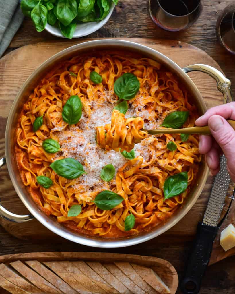 A sauté pan with homemade fresh pasta topped with basil and Parmesan. Someone is twirling a forkful of pasta.
