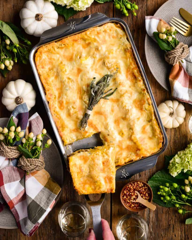 Take a piece out of a squash lasagna in a baking dish. The table is decorated with a fall tablescape.