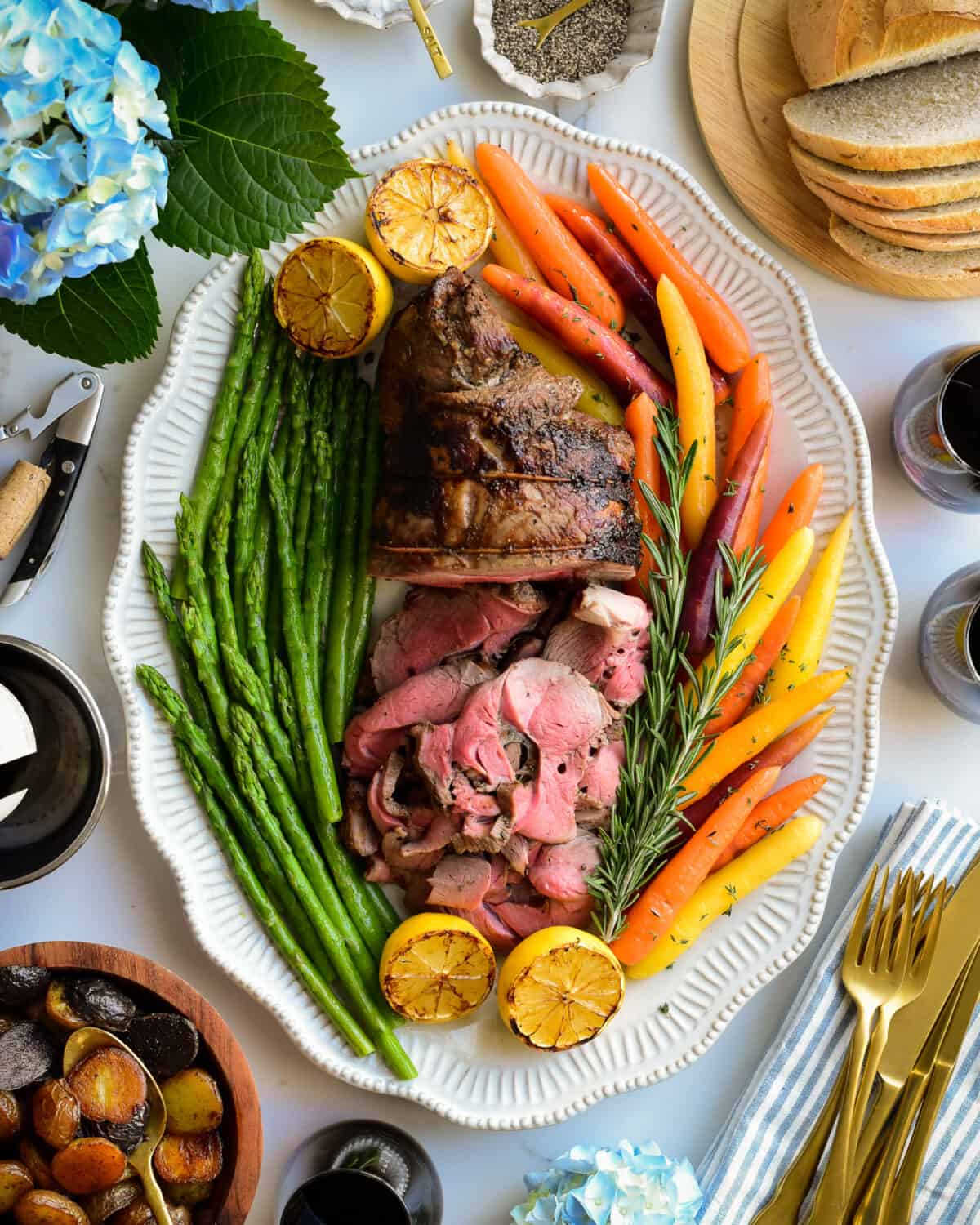 An oval platter with a half sliced rotisserie boneless leg of lamb served with heirloom carrots, asparagus and roasted lemons.