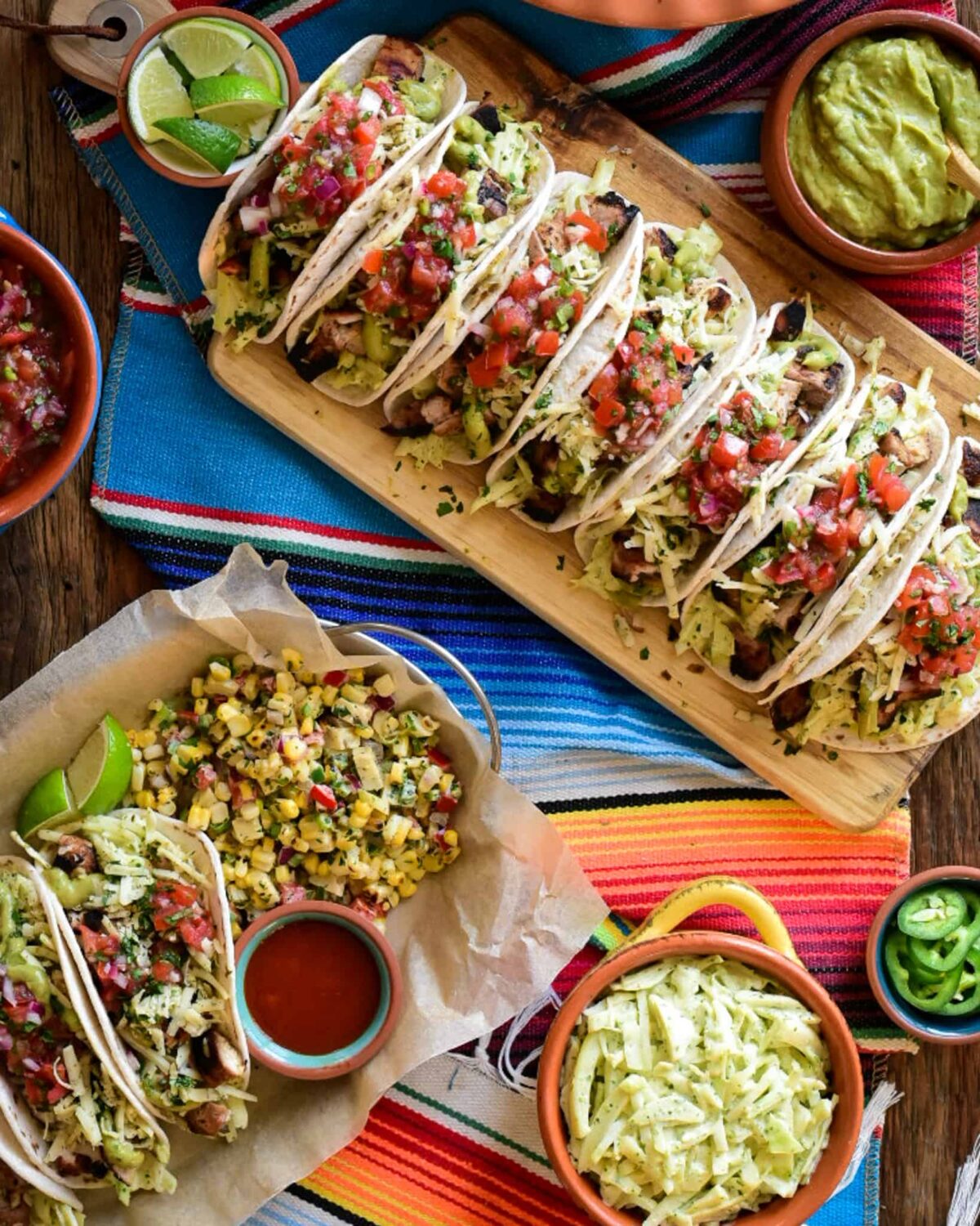 Seven Pollo Asada tacos lined up on a wooden board on a colourful Mexican runner. The tacos are surrounded with dishes of toppings as well as a taco serving with a corn salad. 