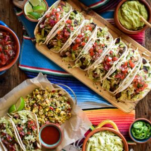 Seven Pollo Asada tacos lined up on a wooden board on a colourful Mexican runner. The tacos are surrounded with dishes of toppings as well as a taco serving with a corn salad.