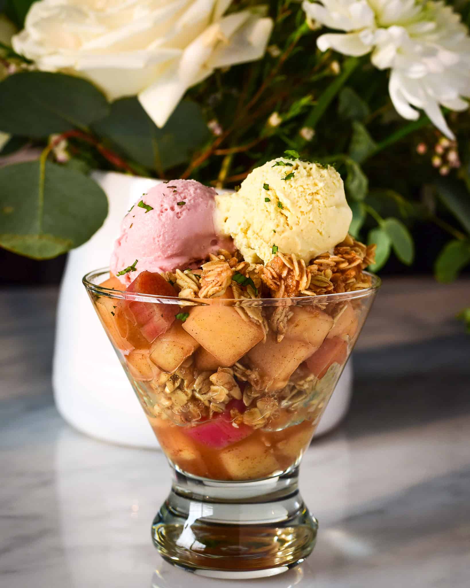 A glass cup filled with layers of apples and rhubarb with a small scoop of vanilla and strawberry ice cream.