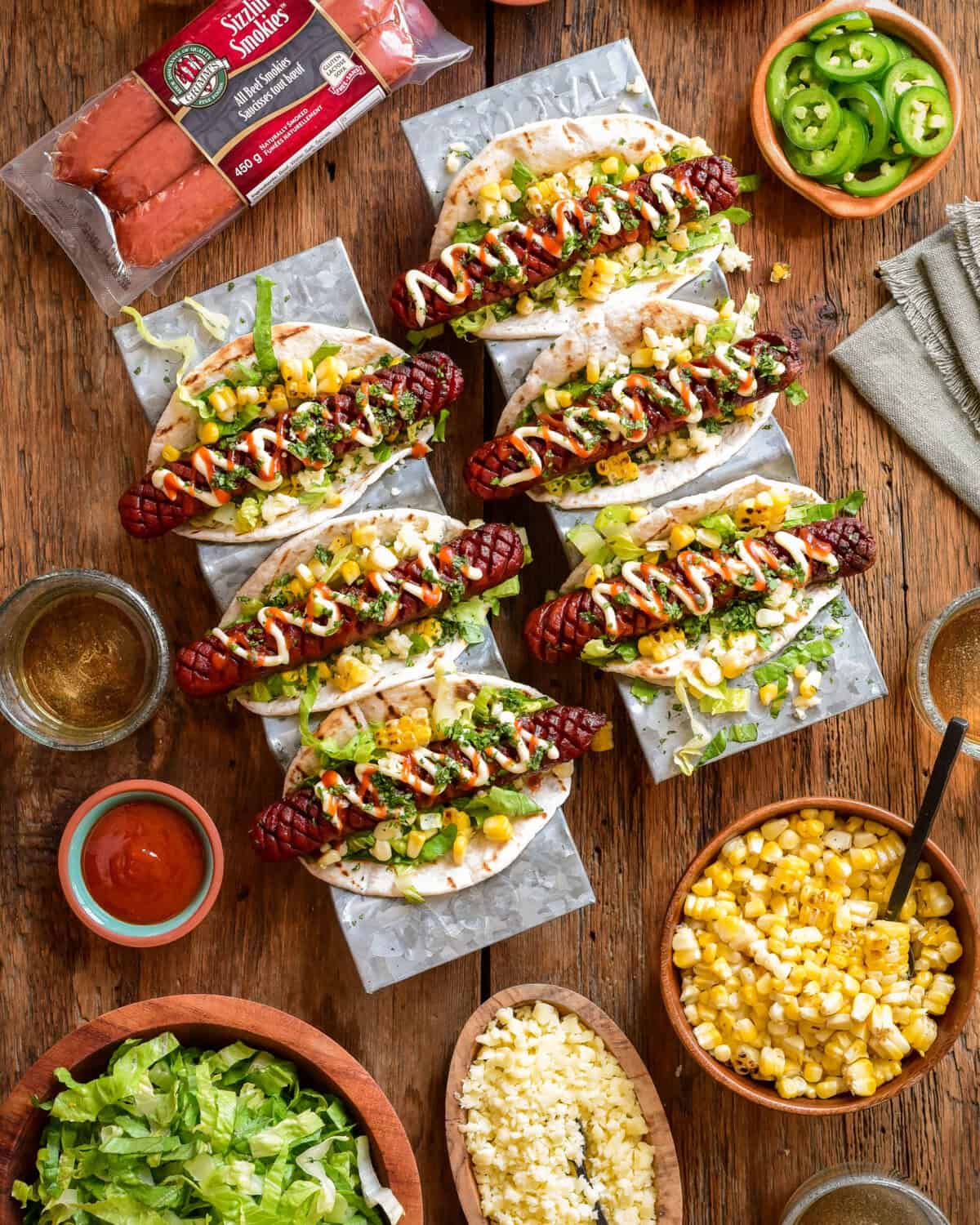 Two taco trays, each filled with three elote-style beef smokies. Extra toppings surround these smokies.