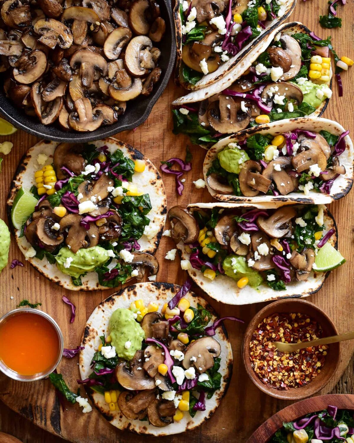 Six Cremini Mushroom Tacos are loaded with a kale-cabbage-corn slaw, crumbled cotija cheese, and a smoky charred jalapeño crema. Alongside a skillet with more sauté mushrooms.
