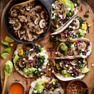 Six Cremini Mushroom Tacos are loaded with a kale-cabbage-corn slaw, crumbled cotija cheese, and a smoky charred jalapeño crema. Alongside a skillet with more sauté mushrooms.